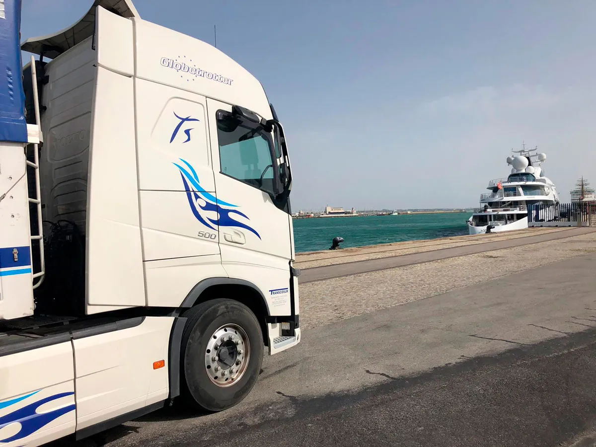 White Transcosur tractor unit with blue livery at a port, with a ship in the background